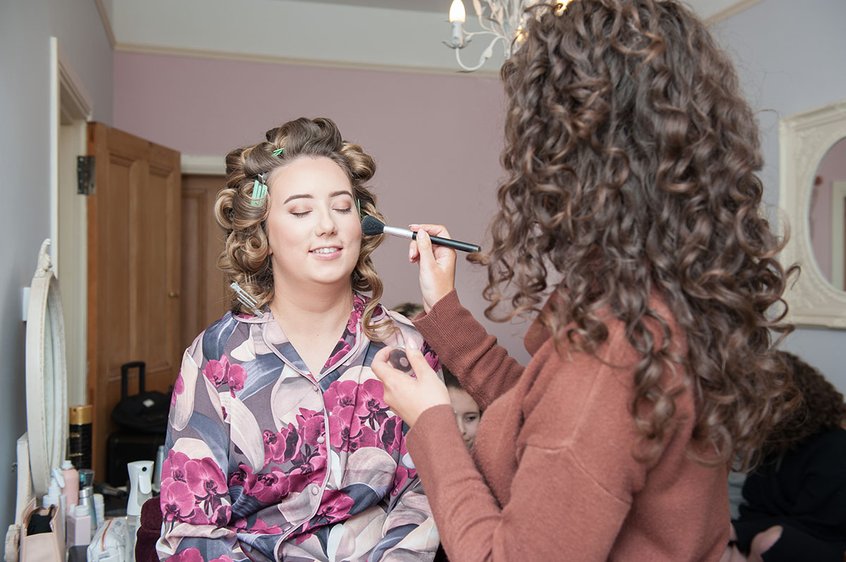 Wedding FAQS - bride wearing flower pyjamas with her hair in curlers, having blusher applied to her cheek by a make-up artist