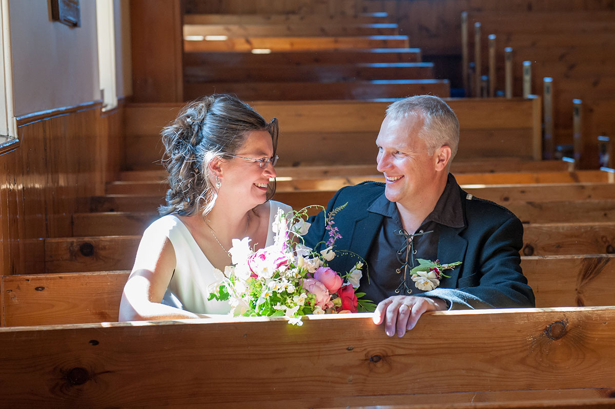 Wedding FAQS - bride and groom sitting in a church pew, facing each other and smiling as the bride holds a bouquet of flowers