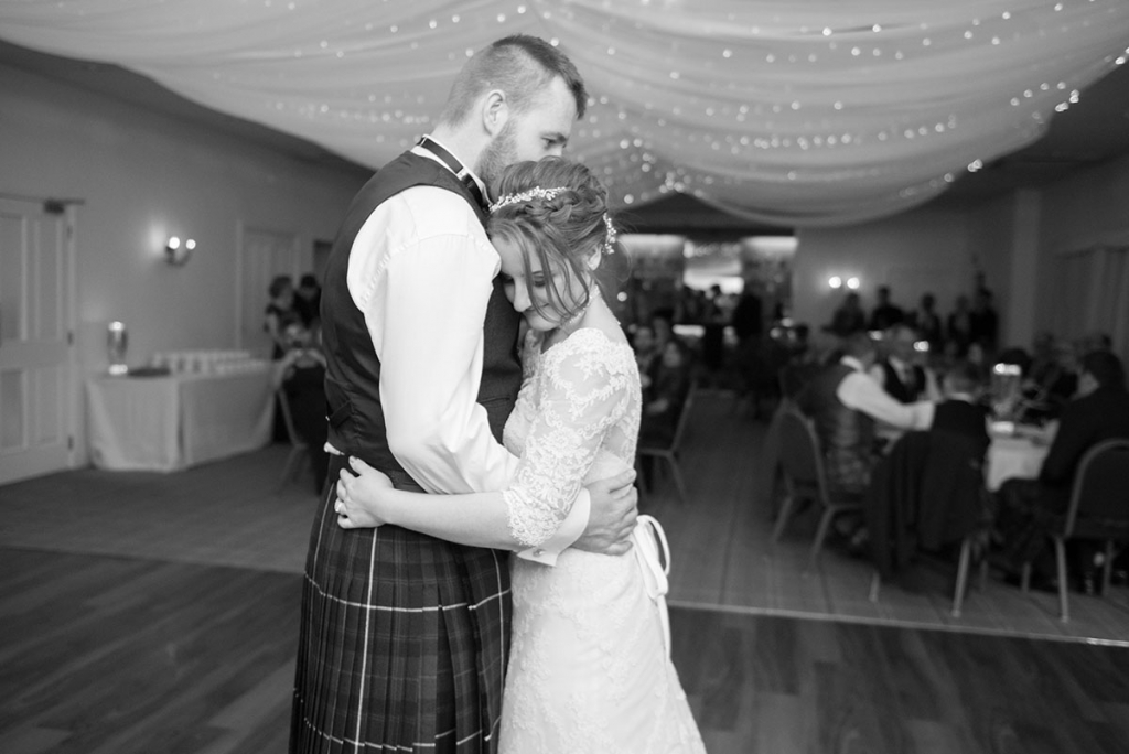 Wedding FAQS - monochrome image of a bride and groom hugging during a dance in a function suite in front of wedding guests
