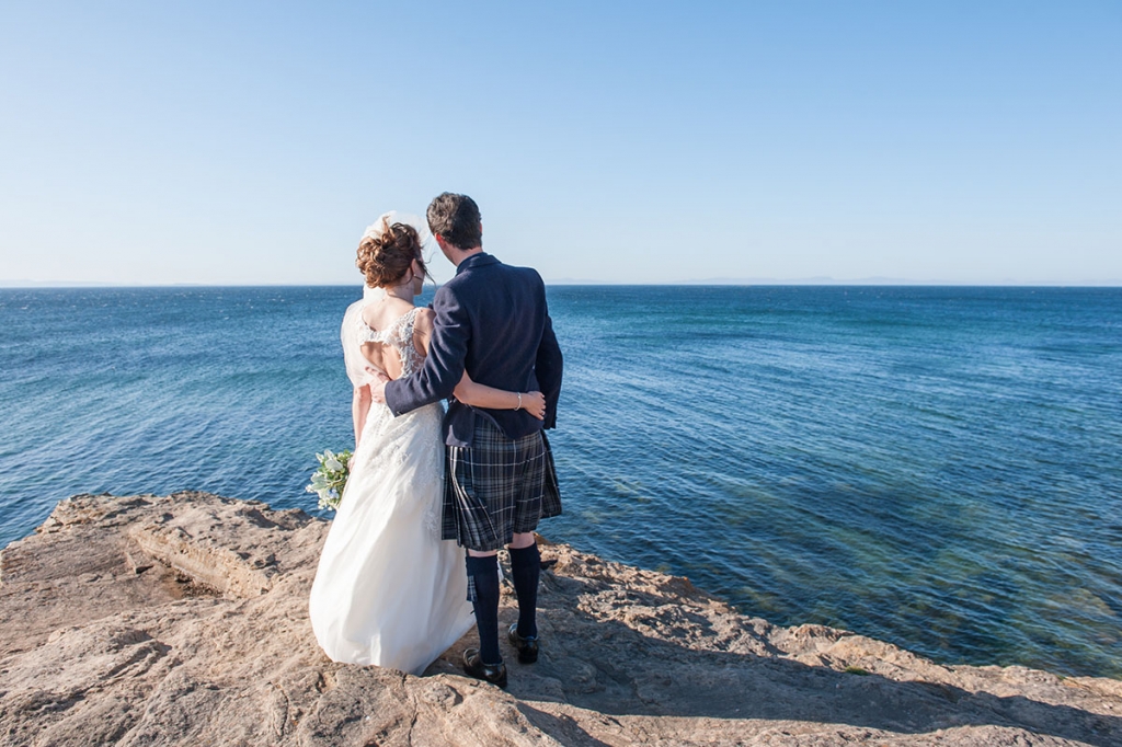 Wedding FAQS - bride and groom standing on a rock with their arms around each other's waists looking out to a blue sea