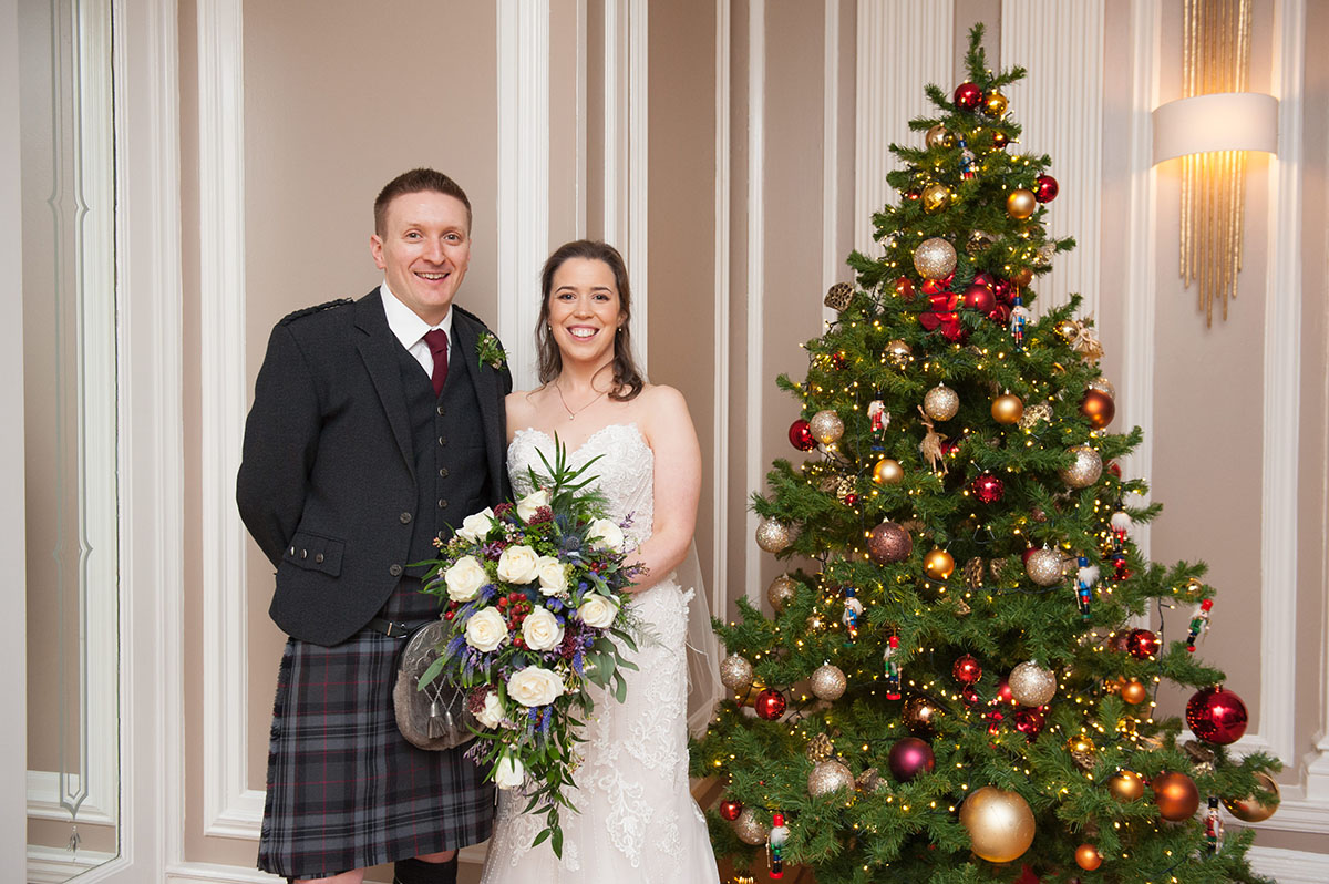 Wedding FAQS - bride and groom standing beside a Christmas tree and with the bride holding a large bouquet of white roses