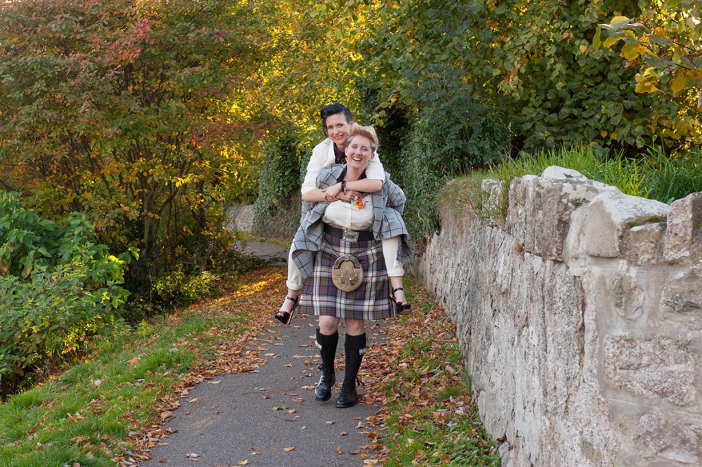 Wedding FAQS - woman dressed in a kilt giving her female partner dressed in a white suit a piggy back next to a stone wall