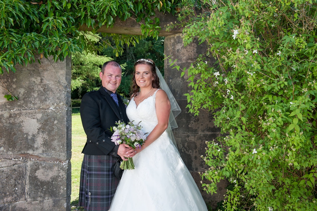 Wedding FAQS - bride and groom with flowers standing in front of a doorway in an outdoor stone wall surrounded by vegetation