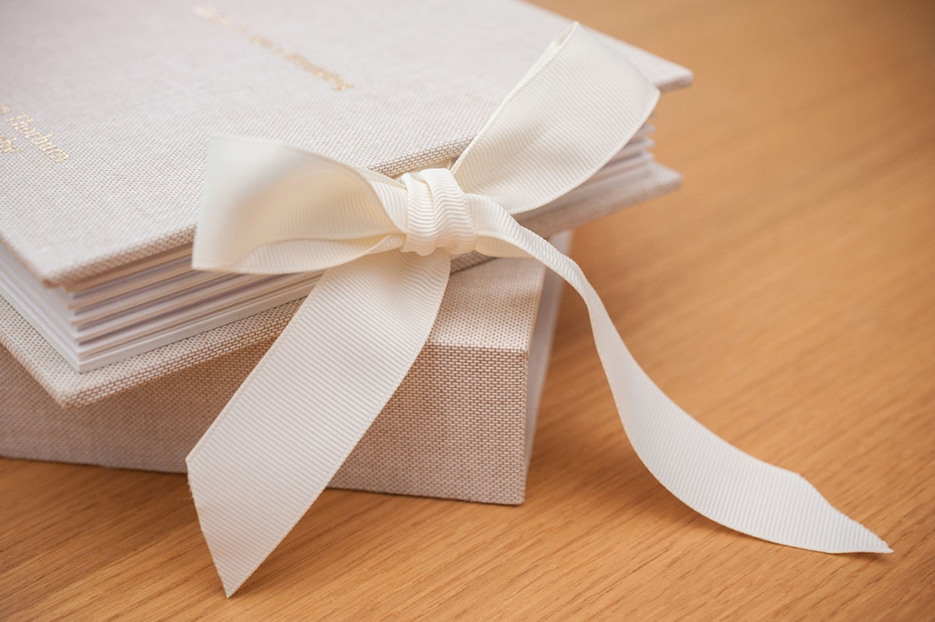 Oatmeal traditional wedding album with cream ribbon tied in a bow, placed on a matching presentation box on a wooden table