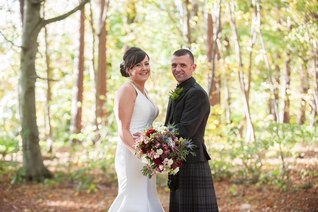 Wedding photograph of a bride and groom, holding flowers, smiling and facing the camera in a woodland
