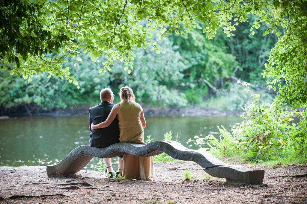 Man and woman sitting on a wooden bench under trees, overlooking a river