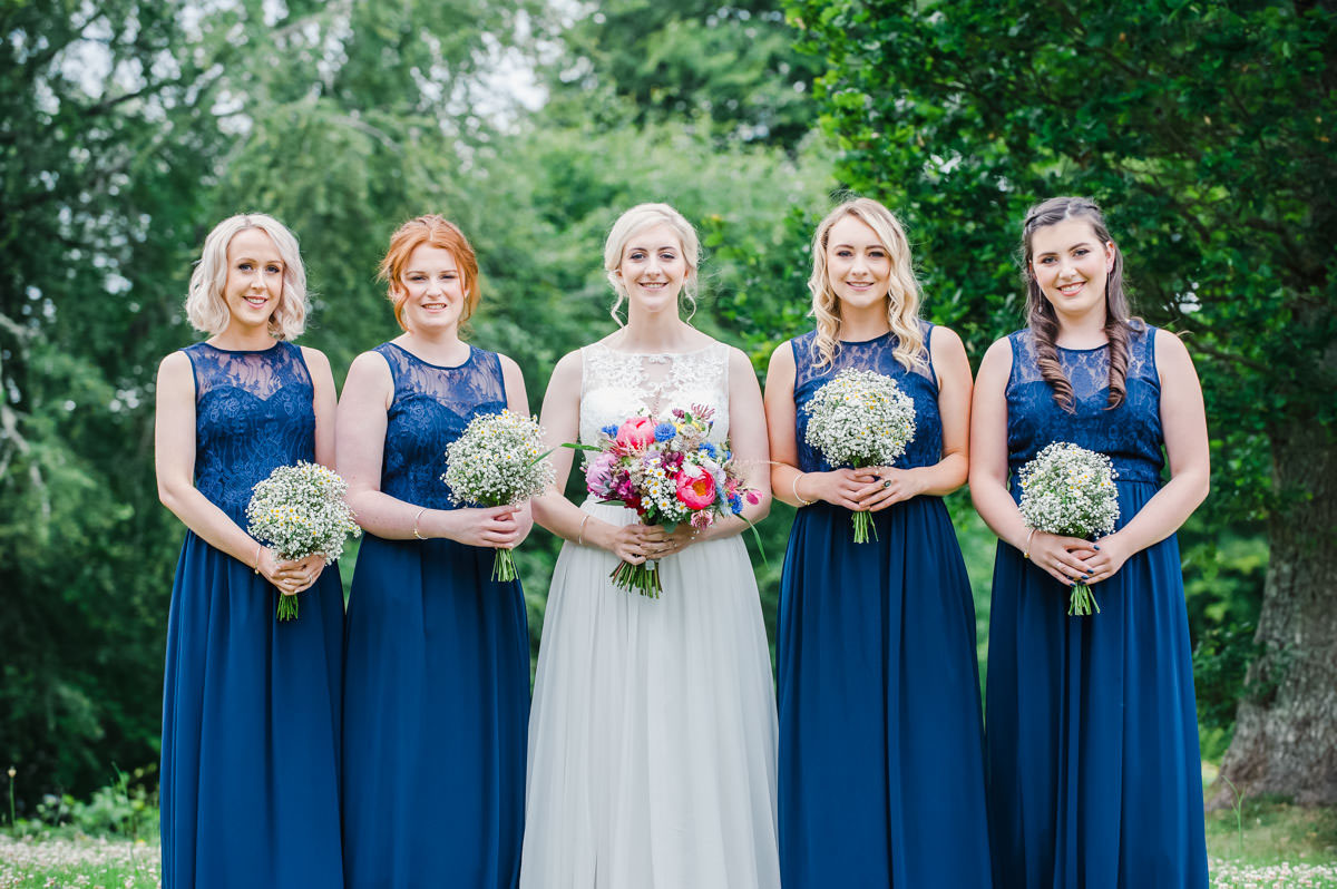 A bride standing in the middle of her four bridesmaids, dressed in navy blue dresses, all holding flowers, in front of trees