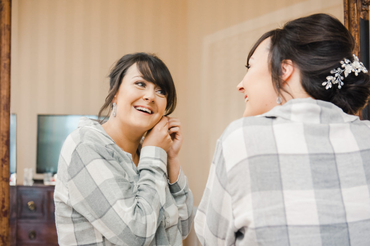 A woman dressed in grey pyjamas looking into a mirror and smiling while inserting an earring into her left earlobe