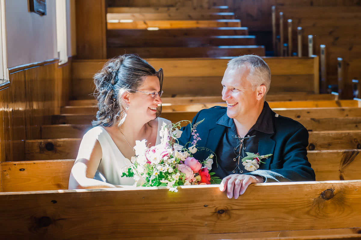 A bride and groom sitting in a church pew, facing each other and smiling as the bride holds a bouquet of flowers