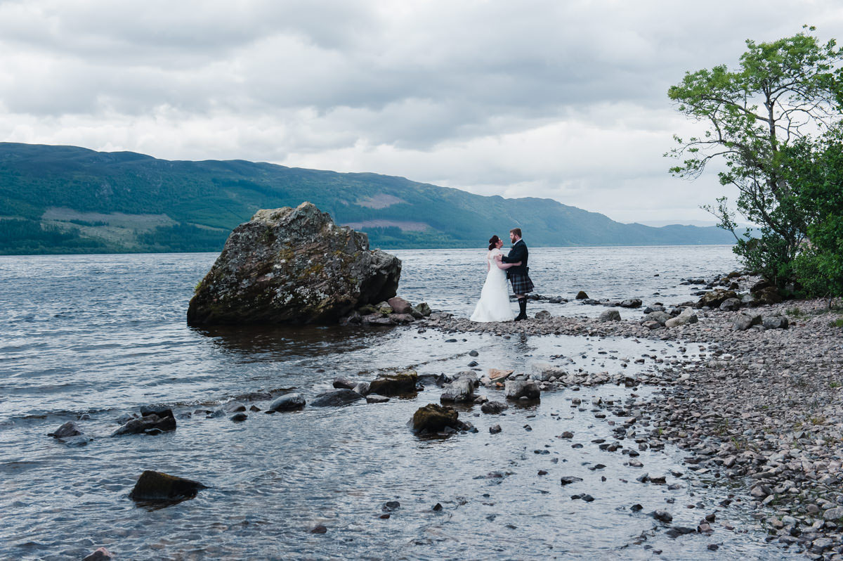 Bride and groom facing each other standing beside a large boulder and tree next to a lake with hills in the background