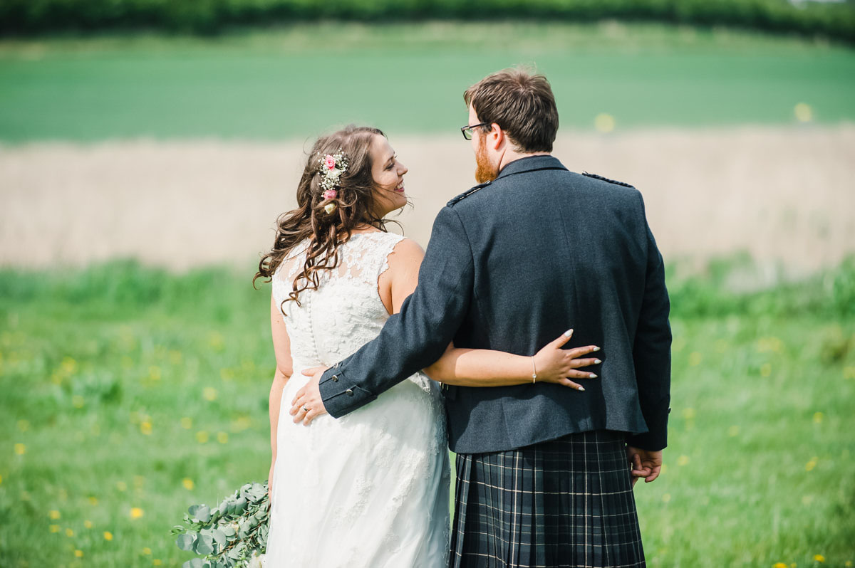 Bride and groom in a green field facing away from the camera with their arms around each other, with the bride smiling