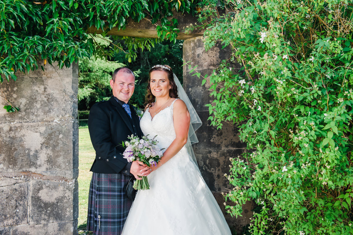 Bride and groom with flowers standing in front of a doorway in an outdoor stone wall surrounded by vegetation