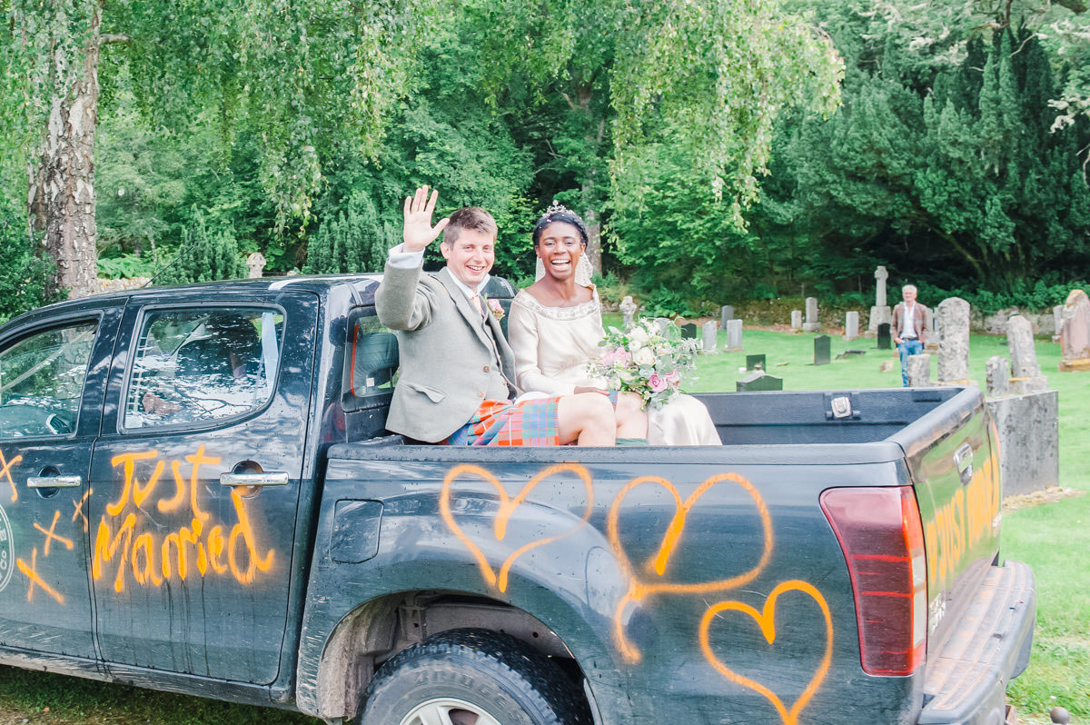 Bride and groom sitting outside on the back of a black truck with orange spray paint that says 'just married'