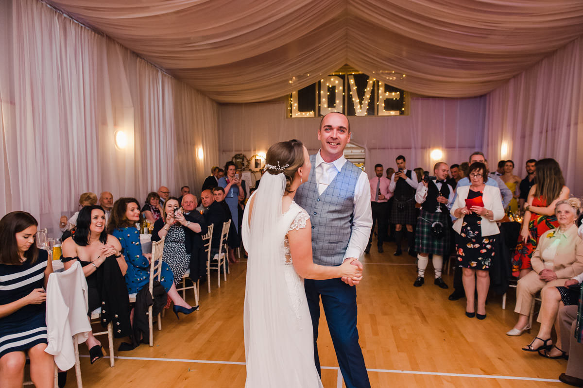 A bride and groom dancing in a village hall in front of wedding guests with the word 'love' lit up in the background