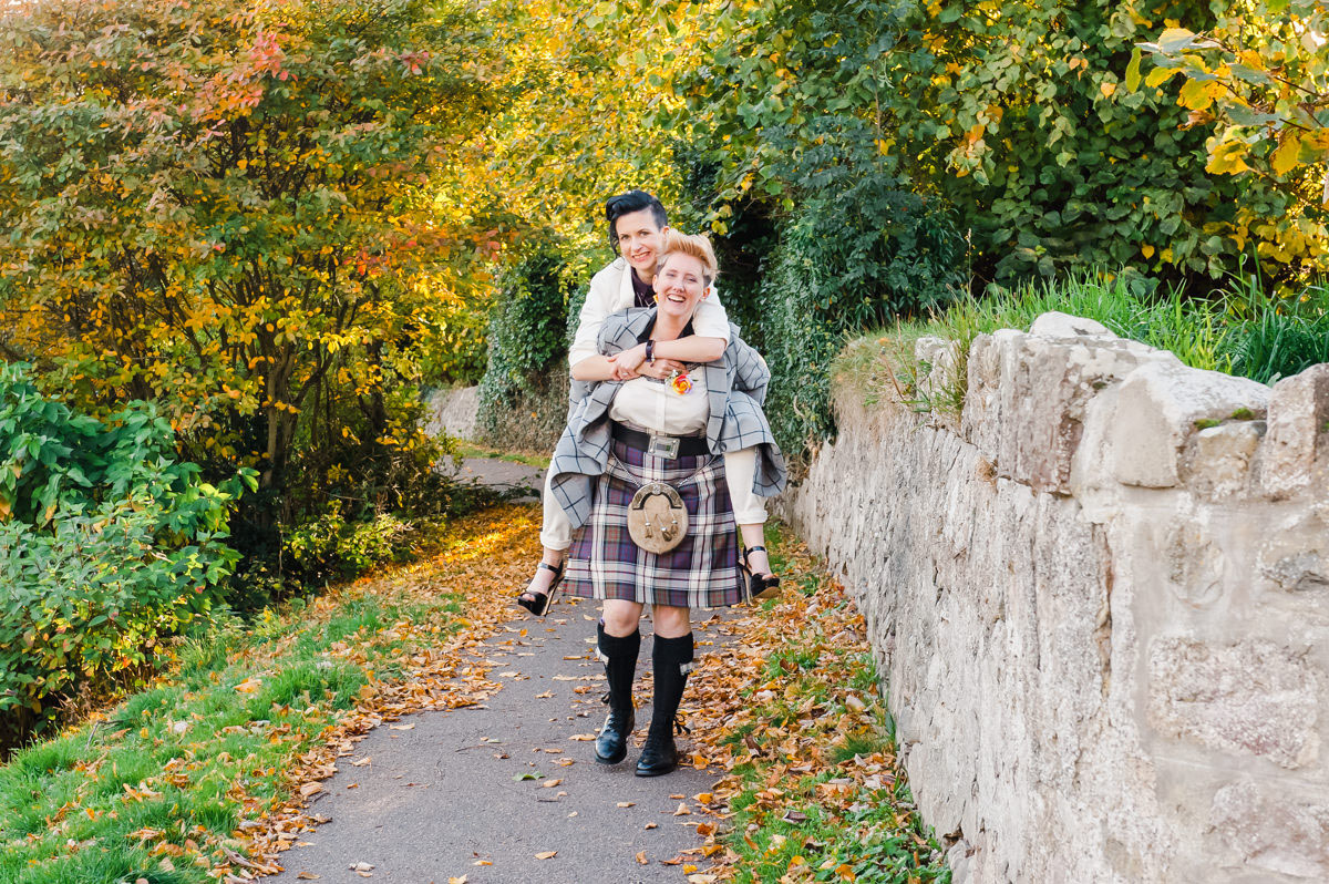Woman dressed in a kilt giving her female partner dressed in a white suit a piggy back next to a stone wall