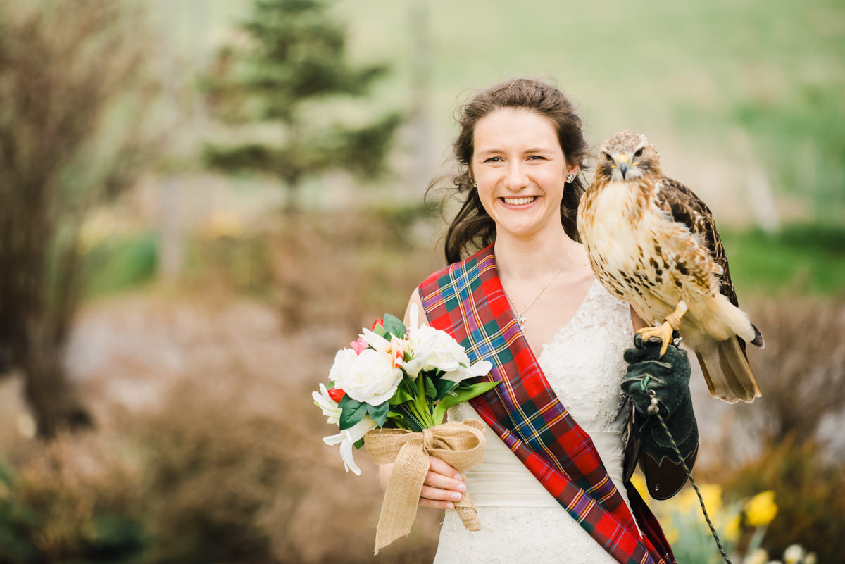 Bride wearing a white dress with a tartan sash holding a bouquet of flowers in one hand, with a bird of prey on the other
