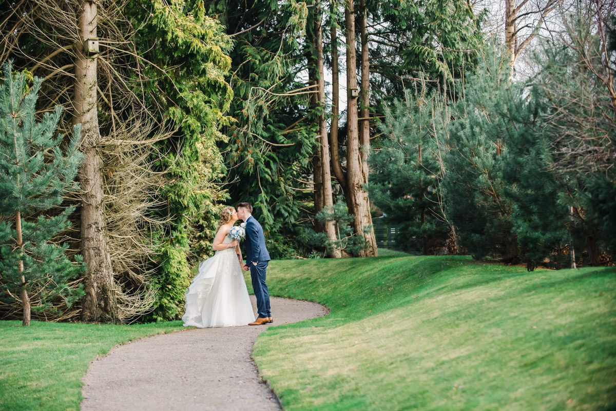 A bride and groom kissing while standing on a path next to grass and underneath trees