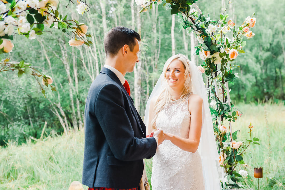 An outdoor wedding ceremony under an arch of flowers, with the bride smiling at her groom