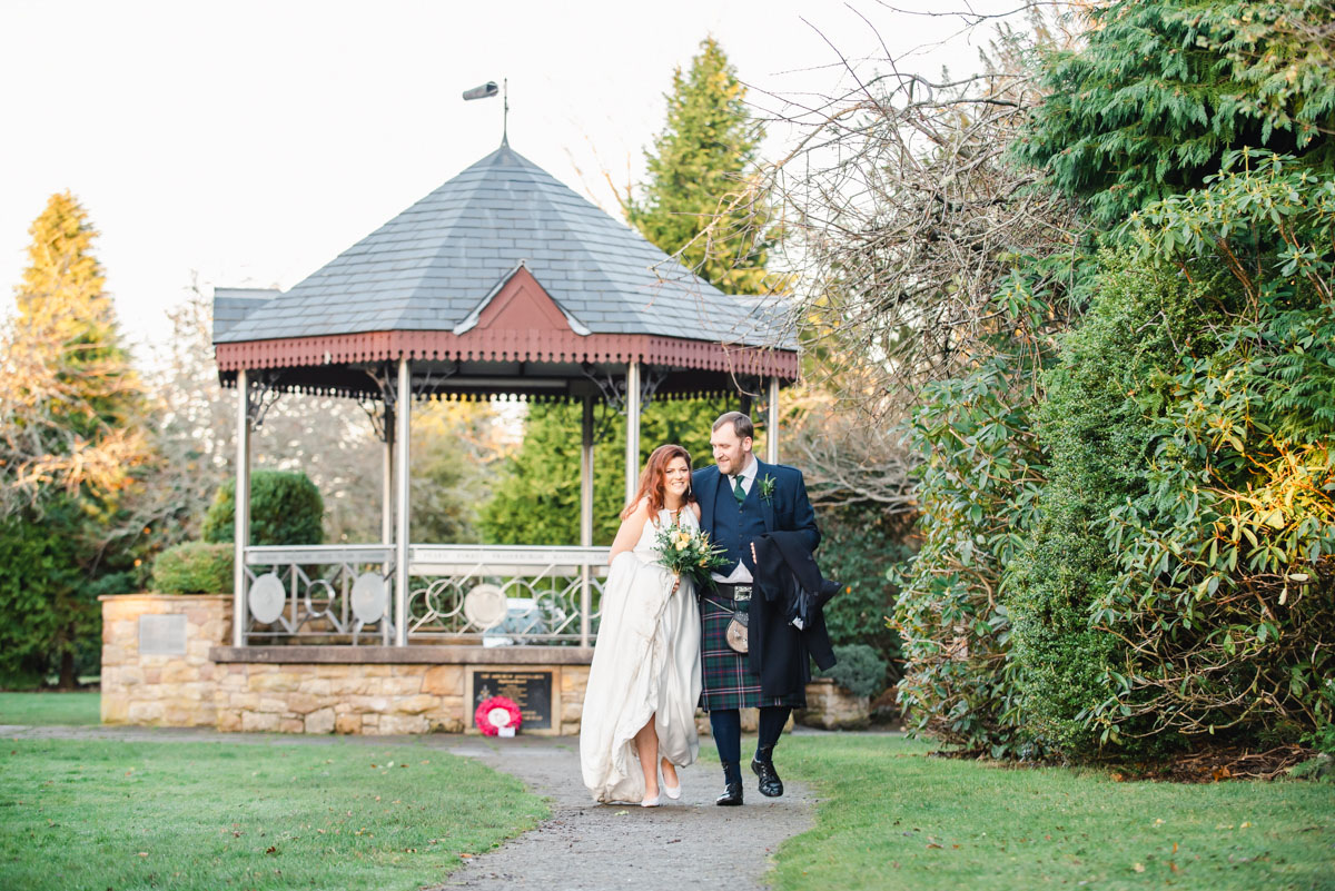 A bride and groom smiling and walking on a path in front of a bandstand in a park with trees