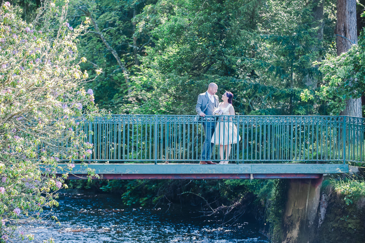 A bride and groom looking at each other, standing on a green footbridge over a river, in front of trees