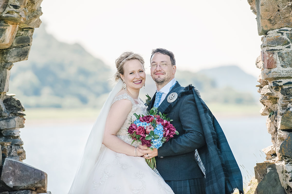 A bride and groom smiling in a stone archway, with water and hills in the distance