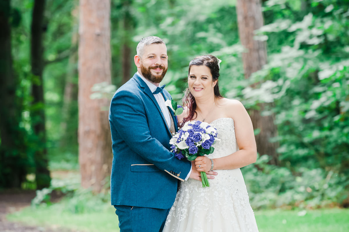 A bride and groom smiling and holding each other in a woodland with mature trees