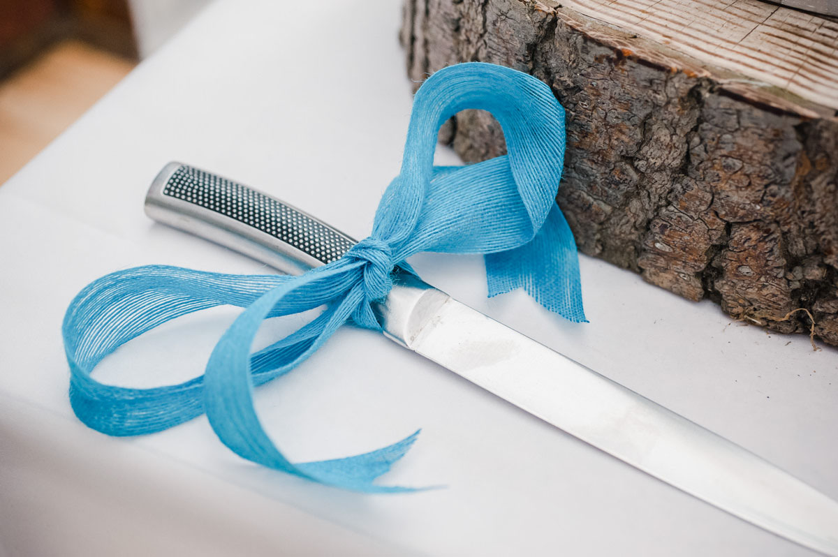 A large knife with a blue ribbon tied in a bow around the handle, placed on a white tablecloth next to a wooden object