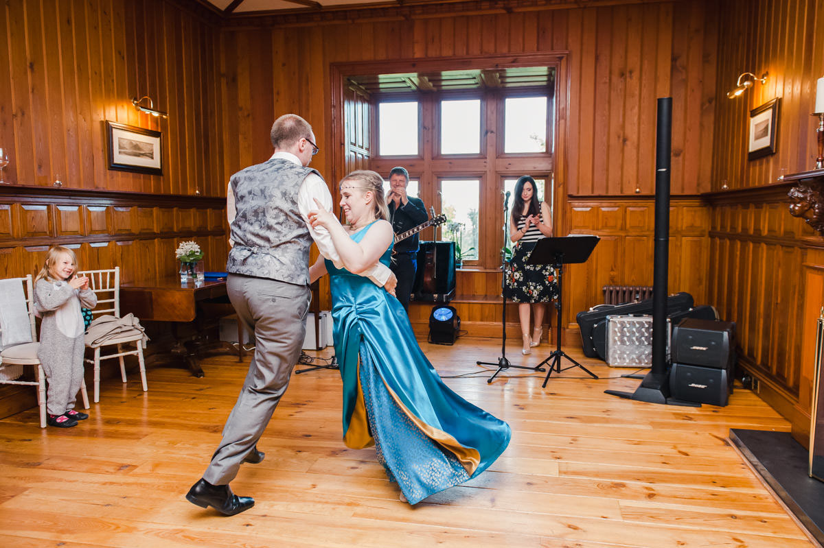A bride in a blue dress dancing with her groom wearing grey trousers and a waistcoat, in front of a two piece wedding band