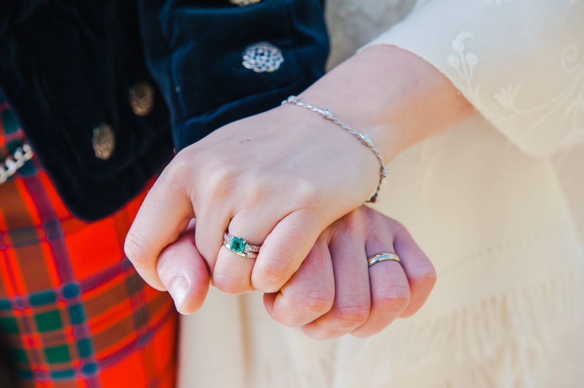 A close-up photograph of a bride and groom holding hands as fists, showing their wedding rings