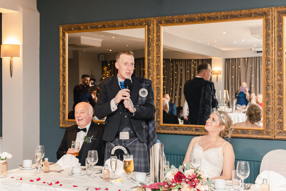 A groom giving his wedding speech standing between the father of the bride and the bride, in front of large mirrors
