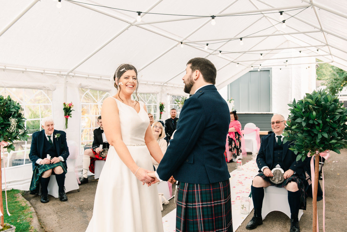 A bride smiling into the camera while holding hands with her groom during her outdoor wedding ceremony in a white marquee