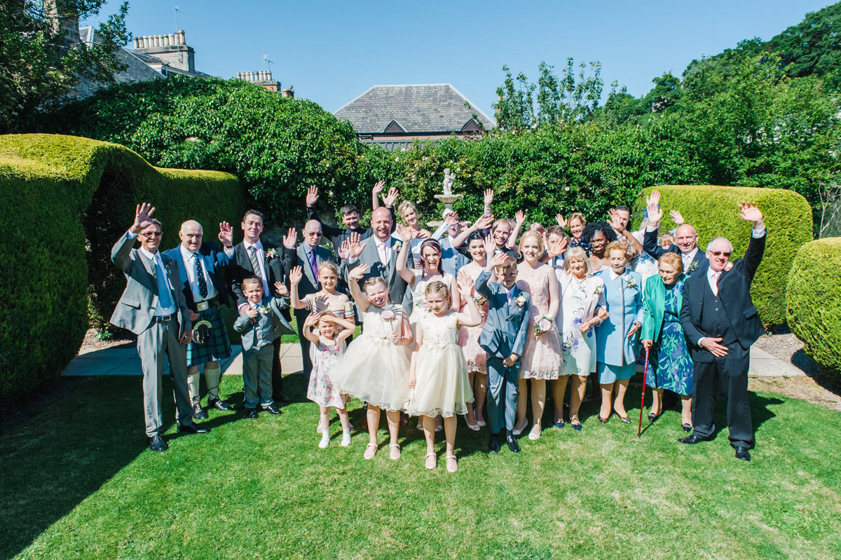 A bride and groom and their wedding guests smiling and waving at the camera standing on a lawn in a garden on a sunny day