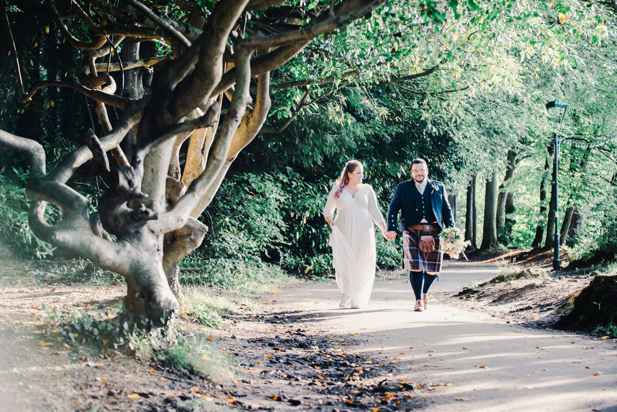 A bride and groom holding hands and walking on a path next to a tree with a twisted trunks in soft sunlight
