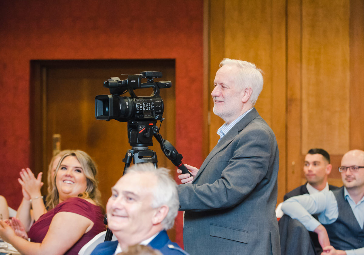 A male wedding videographer dressed in a grey suit, smiling whilst operating his video with guests smiling and laughing