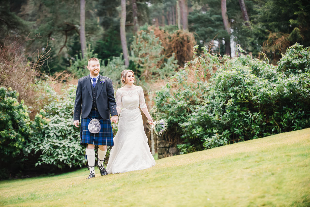 A bride and groom smiling, holding hands and walking on a sloping lawn, with rhododendron bushes and trees in the background