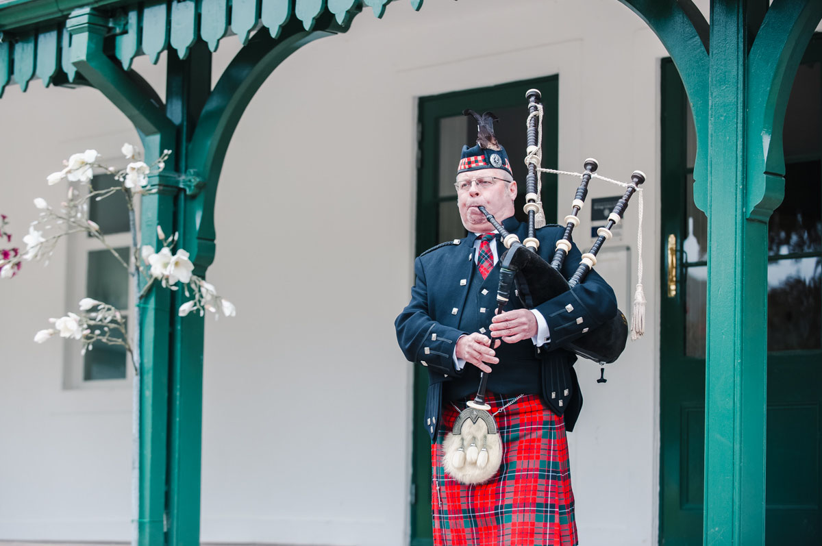 A male piper dressed in a red tartan kilt and tie, and a black jacket and waistcoat. playing the bagpipes in a green archway