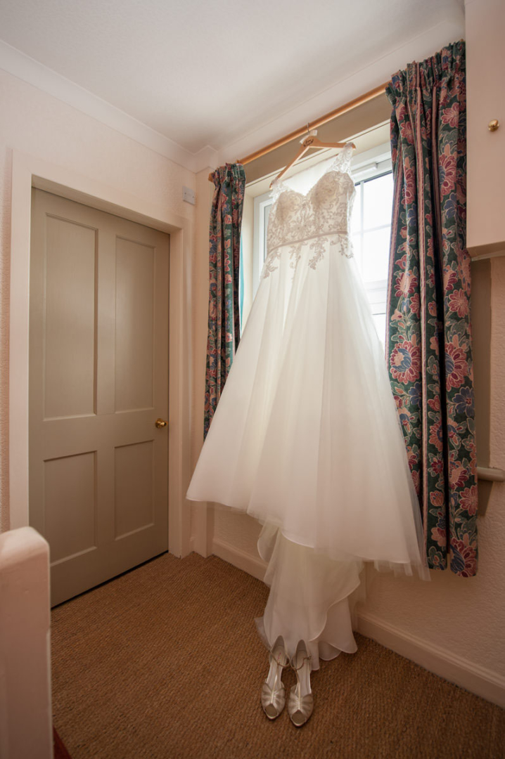 A bride's dress on a coat hanger, hanging in front of a window framed by flower curtains, with the bride's shoes on the floor